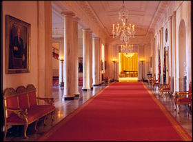 Cross Hall, connecting the State Dining Room and the East Room. To the left is the Entry Hall opening to the North Portico; to the right the Presidential Seal hangs above the entrance to the Blue Room.